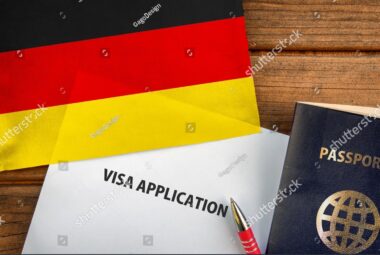 German flag beside a visa application form, passport and red pen on a wooden surface.